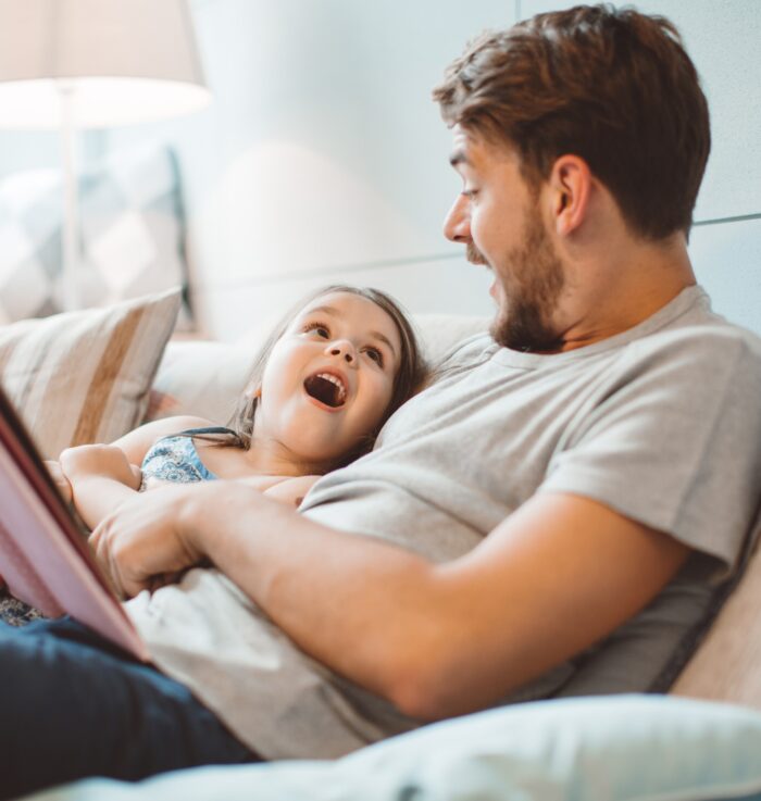 Father and daughter enjoying at home. Sitting on bed and reading book together.